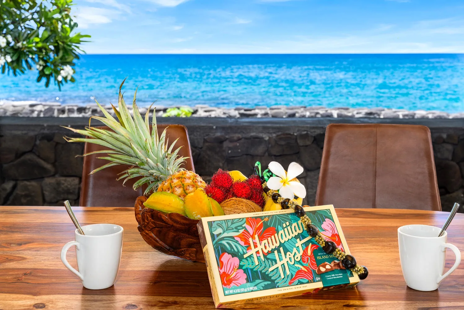 Una mesa en un patio junto al mar muestra fruta y bebidas junto a un muro bajo de piedra.
