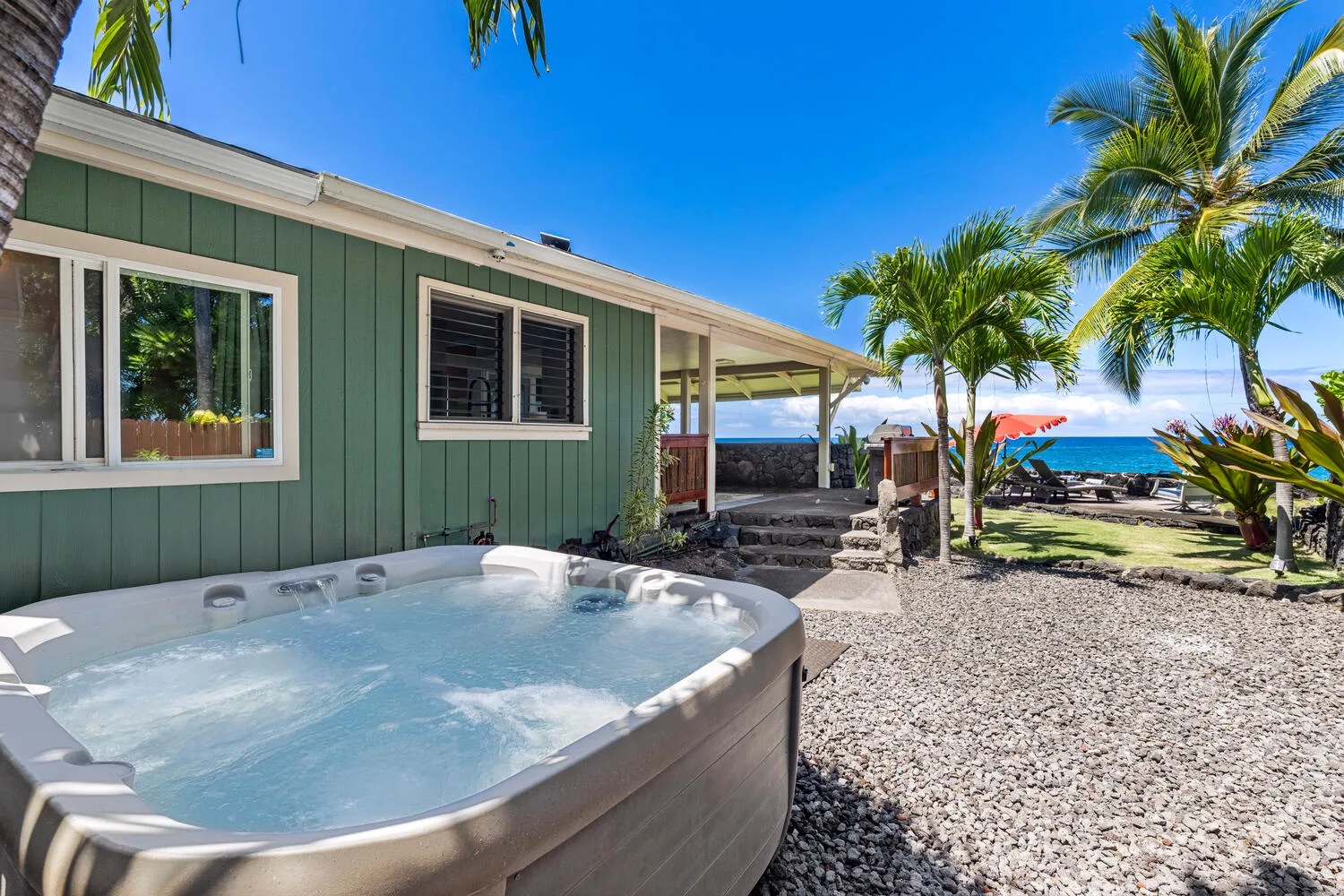 Un jacuzzi está junto a una casa verde, con palmeras y el océano al fondo.
