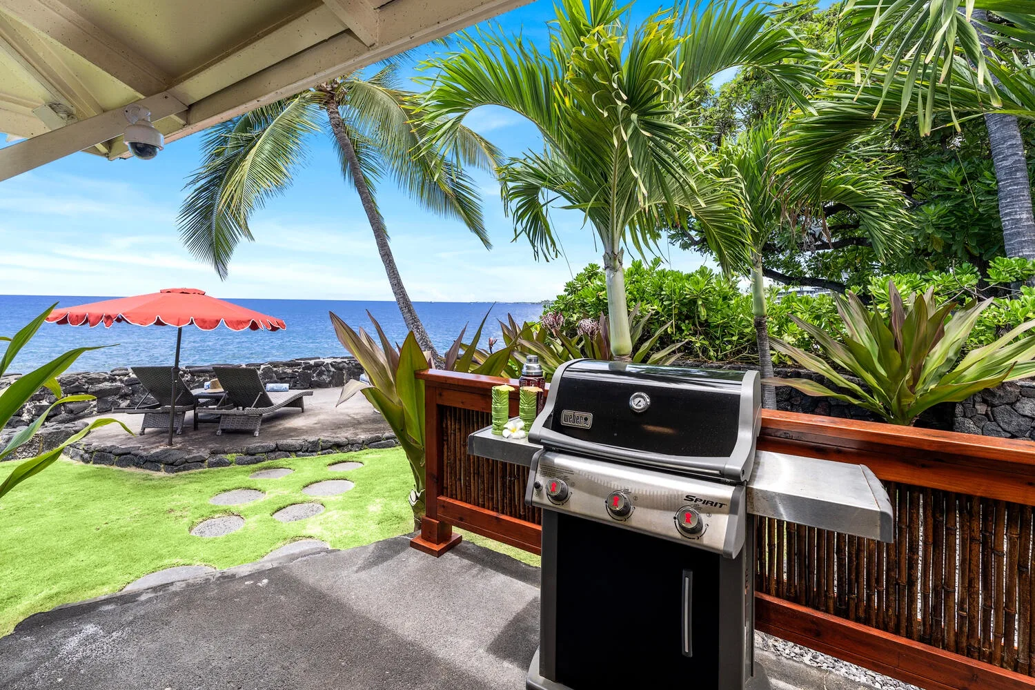 Una parrilla y un patio al aire libre dan a una costa tropical con palmeras y tumbonas.