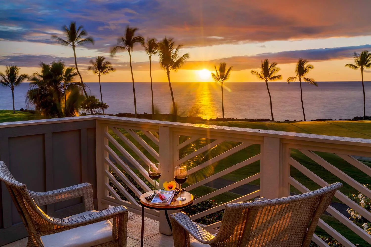 A balcony seating area overlooks palm trees and the ocean at sunset.