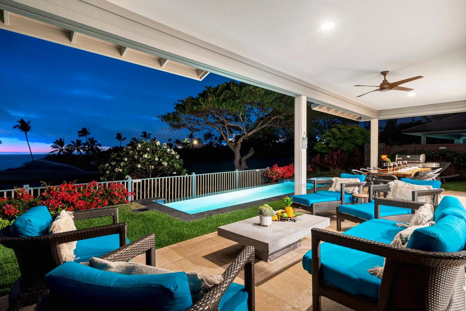 A covered outdoor patio with turquoise seating overlooks a pool and tropical garden at dusk.