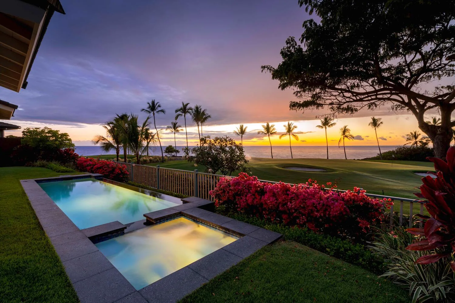 Una piscina y un jacuzzi dan a un césped, palmeras y el mar al atardecer.