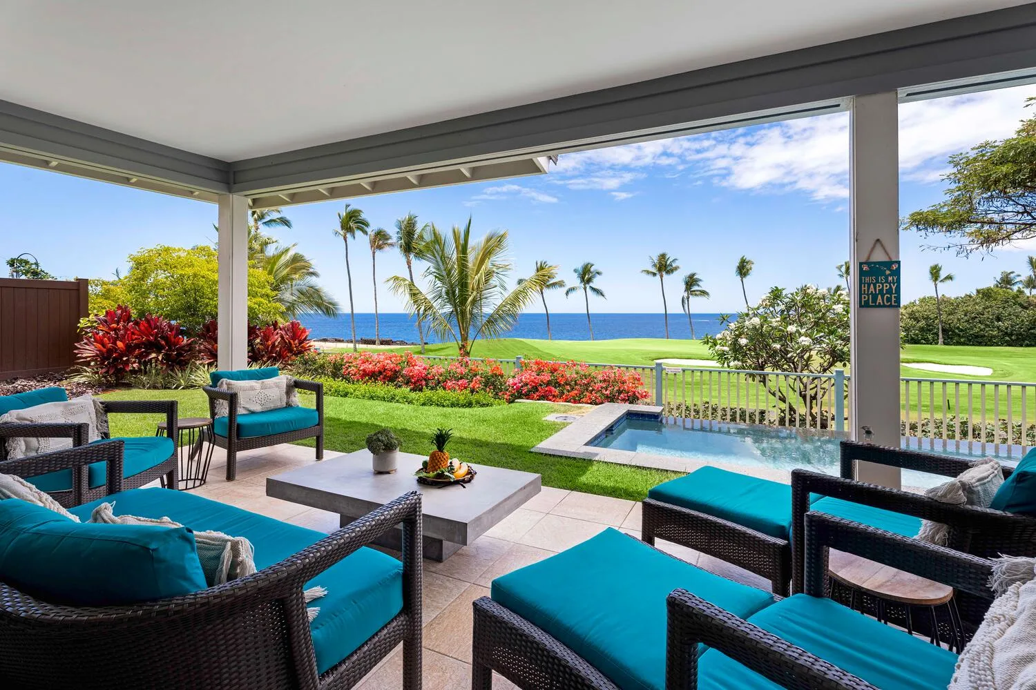 A covered patio with cushioned seating overlooks a garden, pool, and ocean.