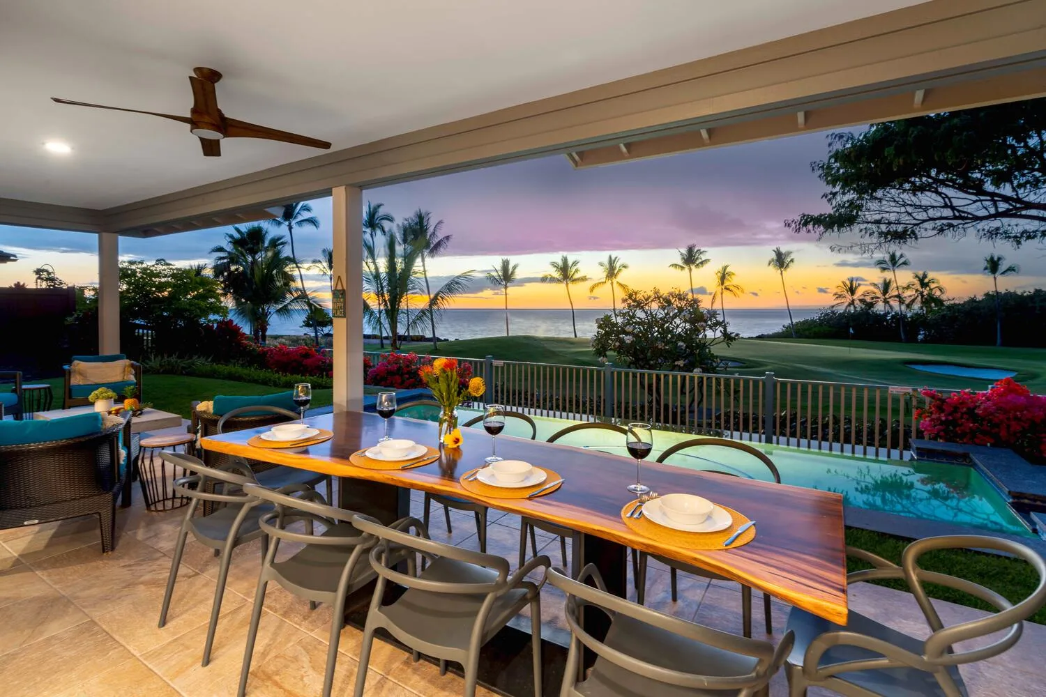 Un comedor exterior cubierto tiene vistas a una piscina y al océano bordeado de palmeras al atardecer.