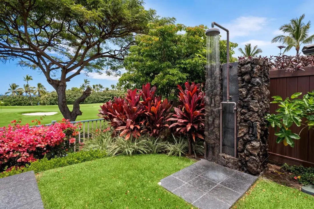 A garden shower stands beside tropical plants and a green lawn.