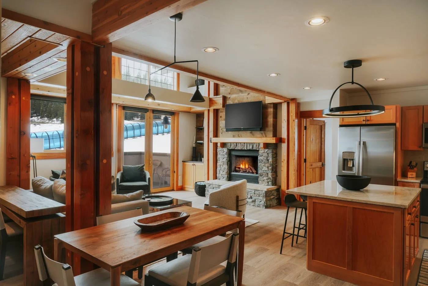 A wood-paneled living room and kitchen with a stone fireplace and large windows.