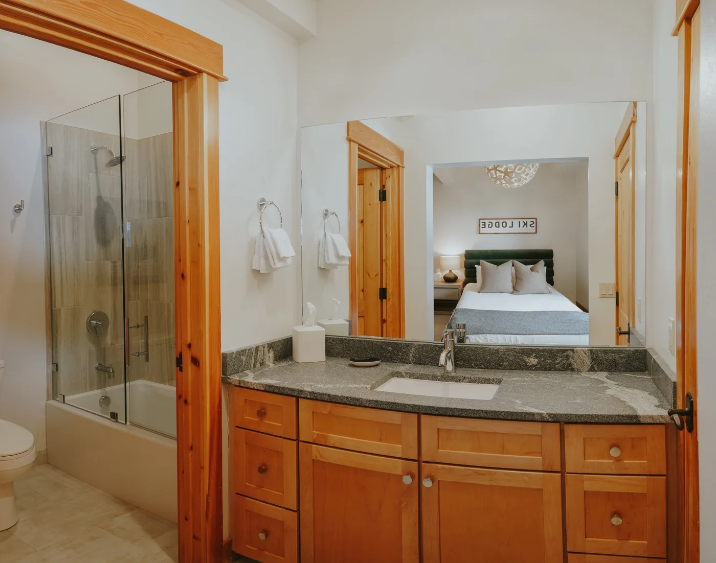 Un lavabo de baño está junto a una ducha de vidrio, con un dormitorio reflejado en el espejo.