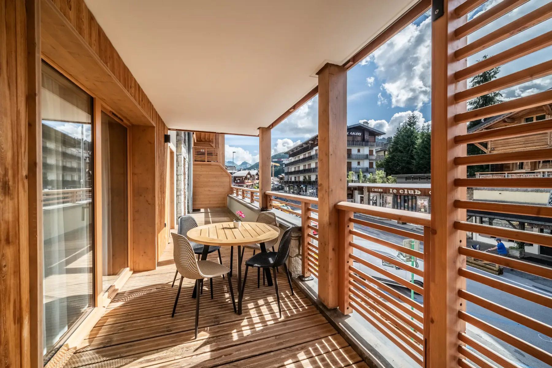 A wooden balcony with outdoor seating overlooks nearby buildings and mountains.