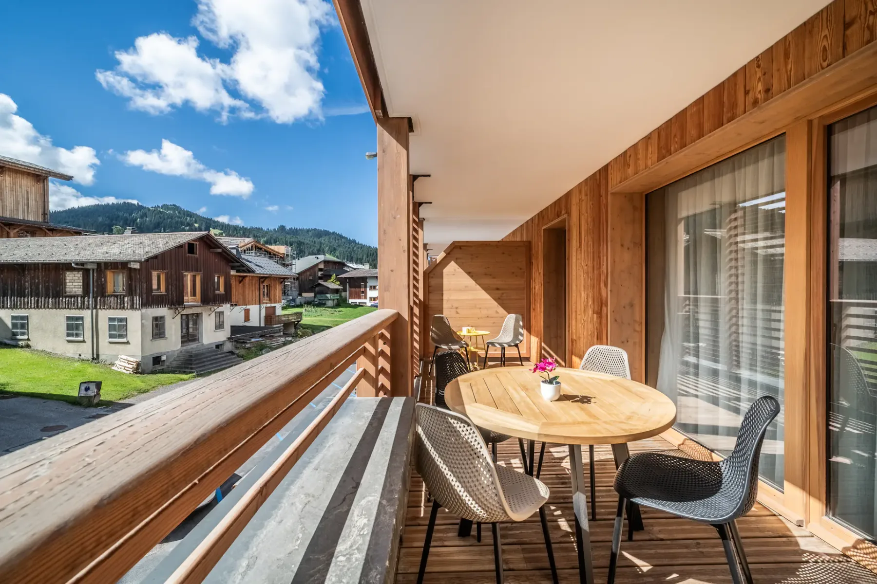 A wooden terrace has an outdoor table and seating overlooking nearby buildings and mountains.