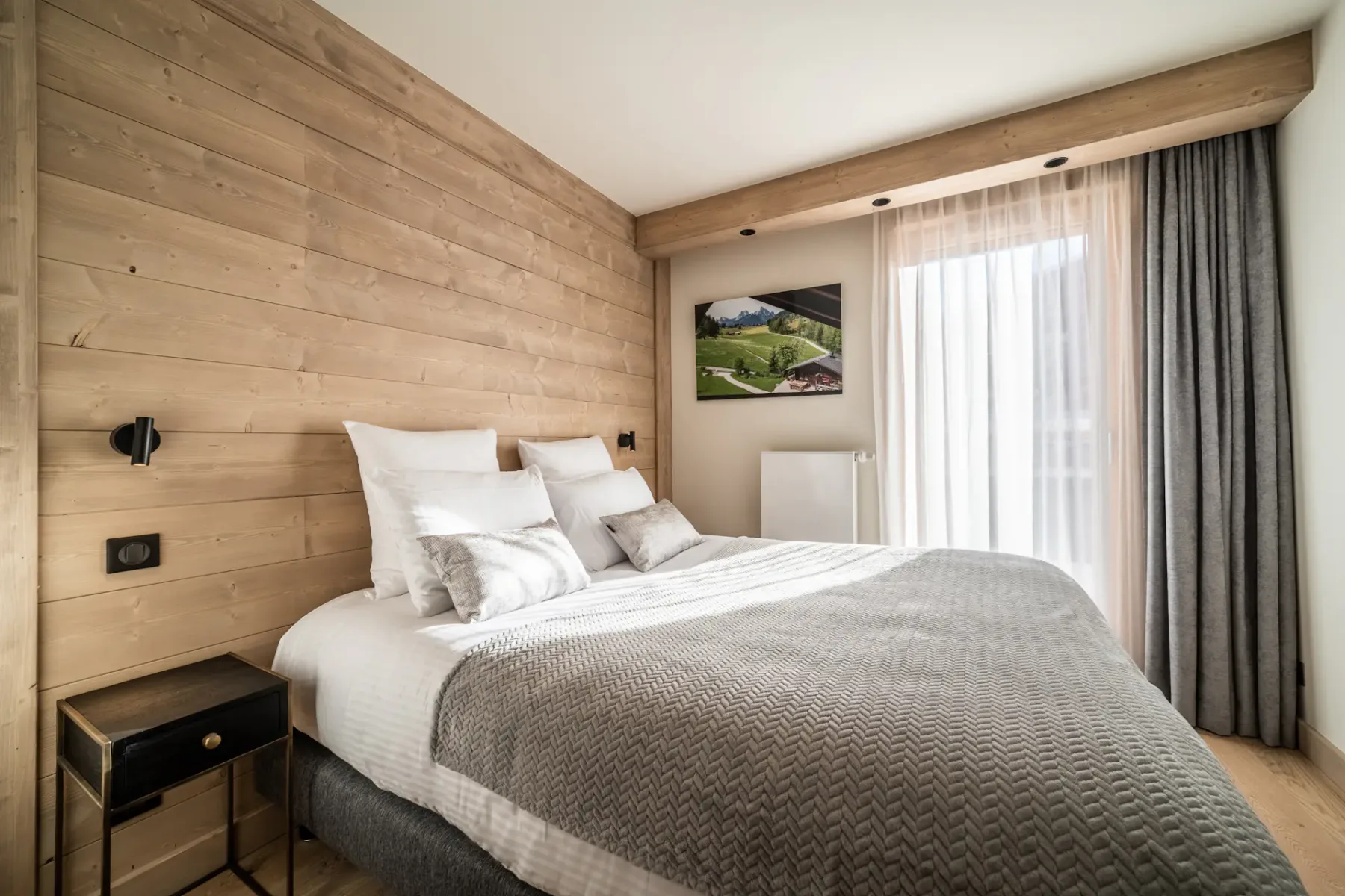 A bedroom with a neatly made bed beside a large curtained window.