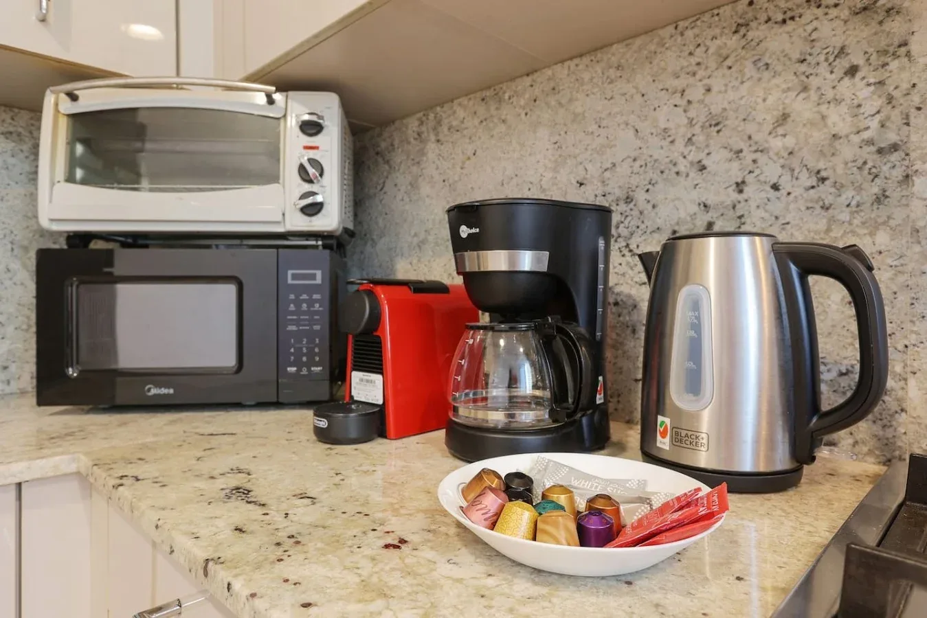 Una encimera de cocina con microondas, cafetera, hervidor y un cuenco con productos para té.