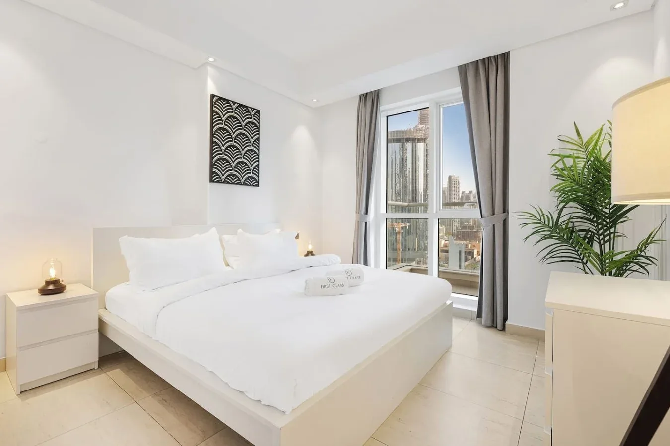 Un dormitorio luminoso con una cama blanca, ventanas altas y cortinas grises.