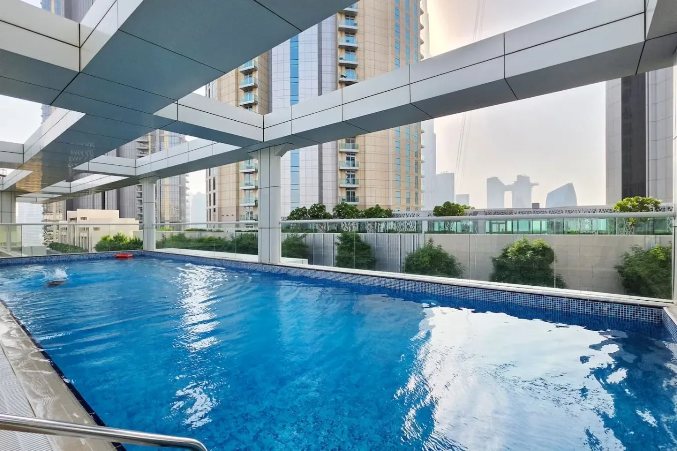 Una piscina al aire libre se encuentra bajo una estructura moderna, con edificios altos al fondo.