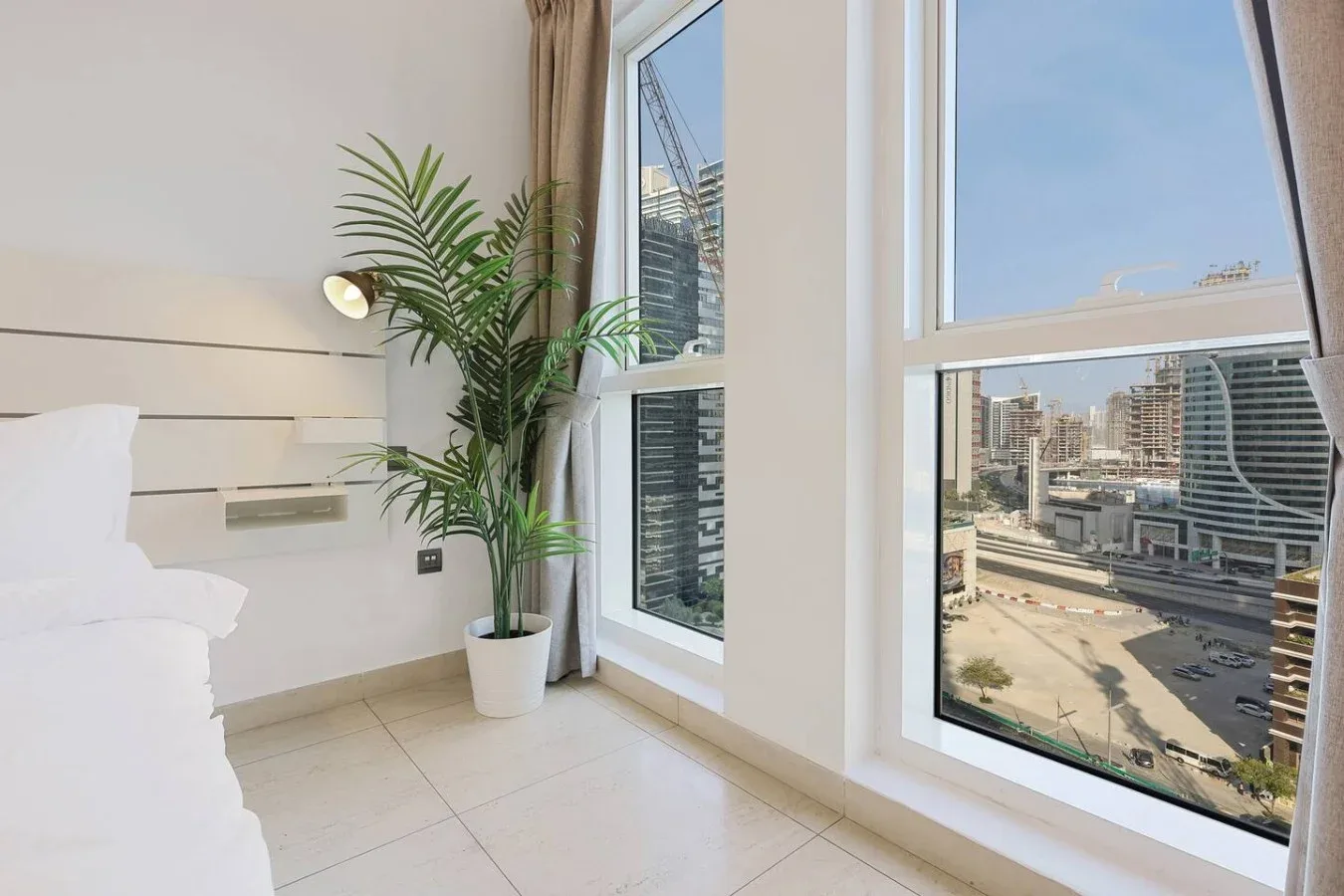 Un dormitorio luminoso con una cama, una planta en maceta y grandes ventanas con vistas a la ciudad.
