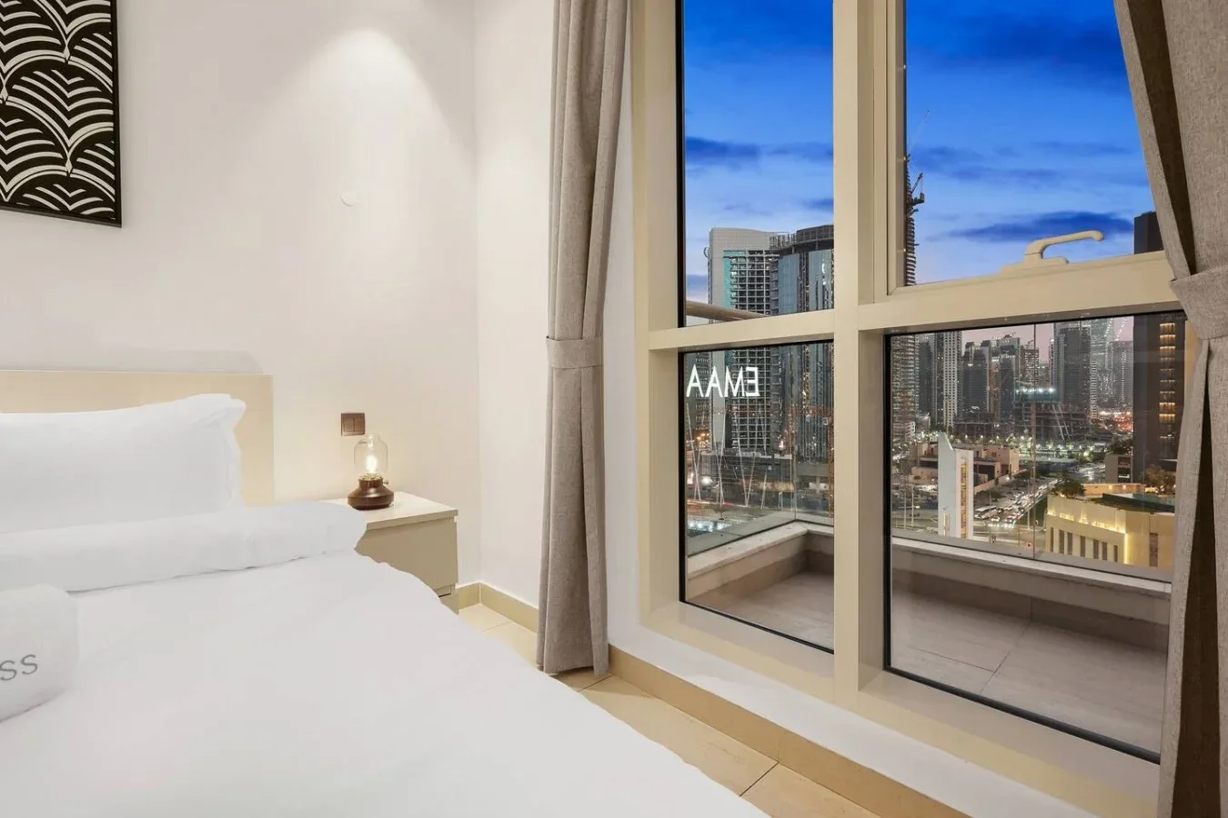 Dormitorio con ropa de cama blanca junto a grandes ventanas con vistas a la ciudad.