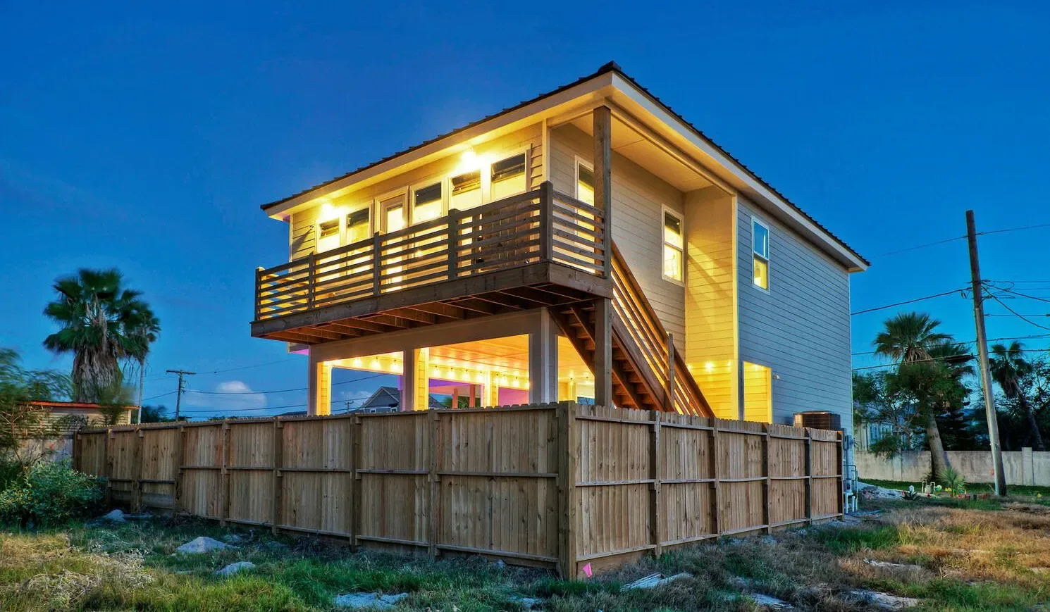 A raised wooden house with a balcony stands behind a fenced yard at dusk.