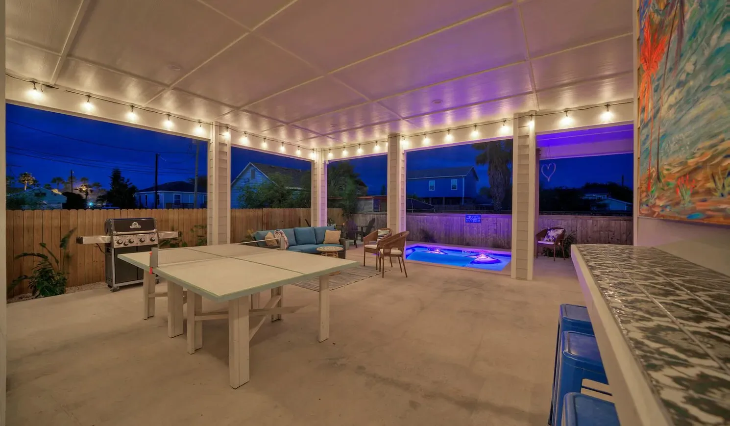 A covered patio dining area with a table, grill, and illuminated swimming pool.