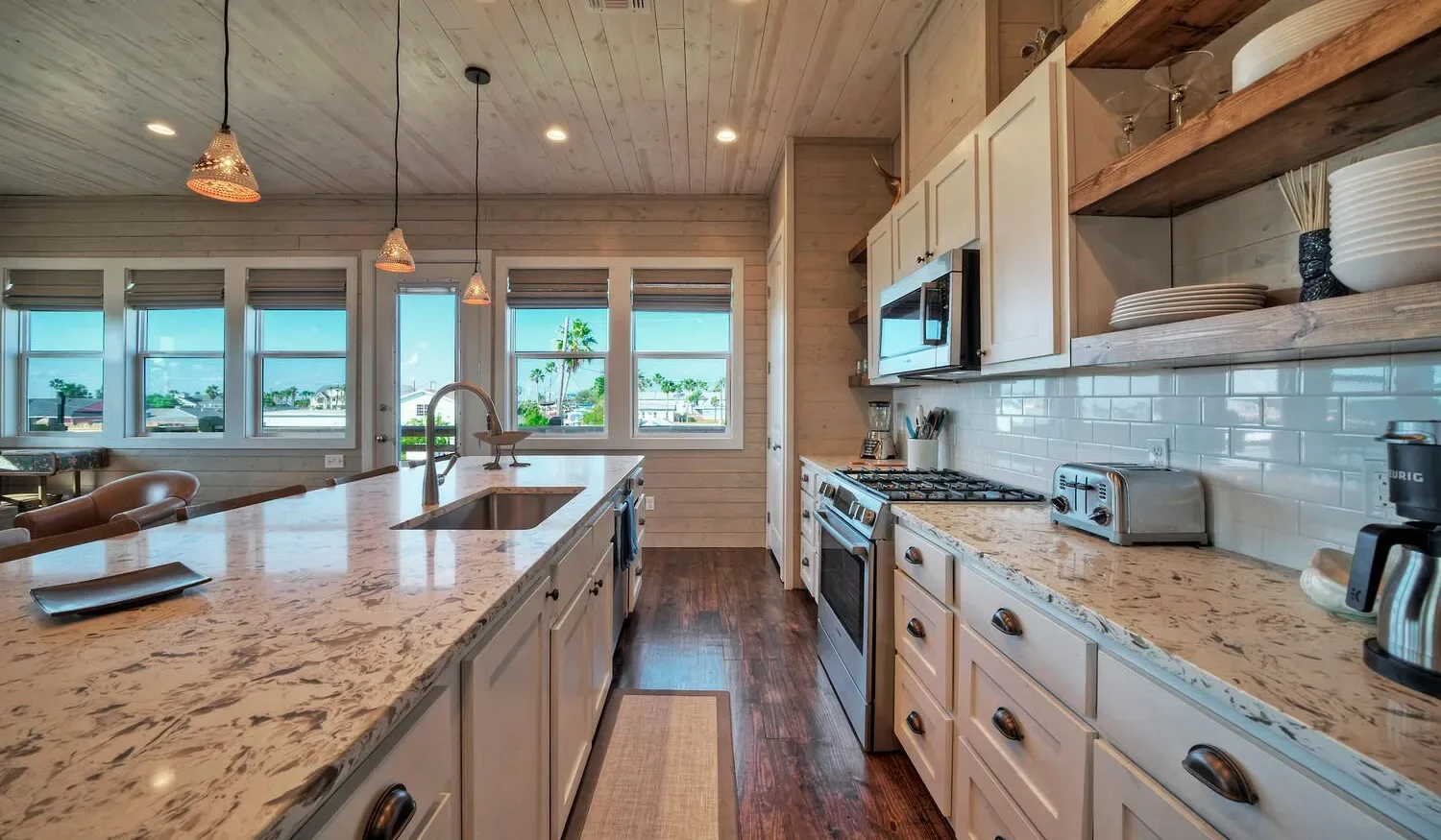 A bright kitchen with a long island, pendant lights, and windows overlooking the water.