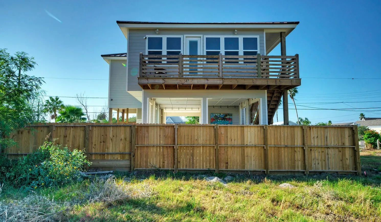 A raised house with a wooden balcony stands behind a wooden fence.