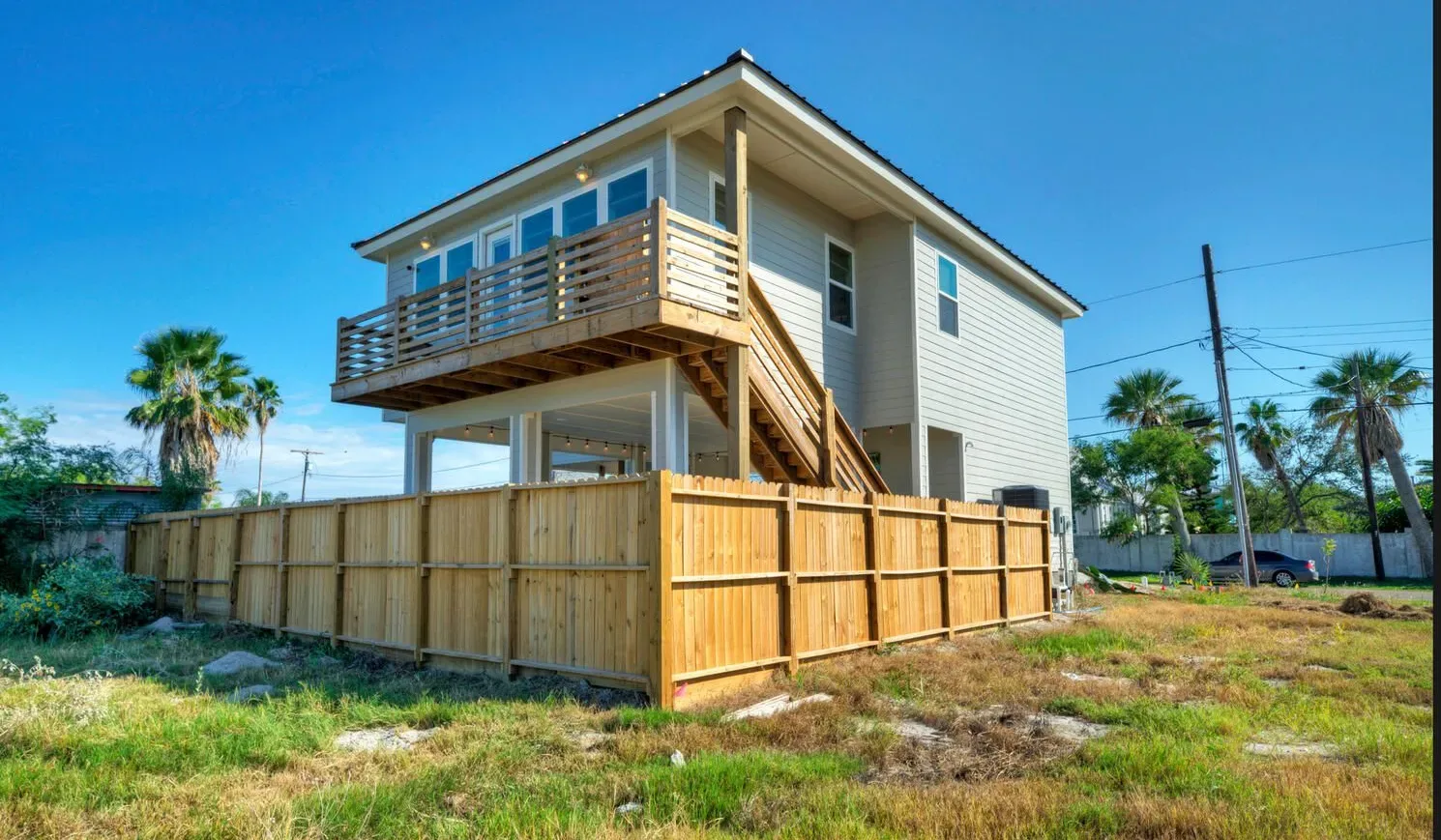 A raised house with a wooden deck and fenced yard under a clear blue sky.