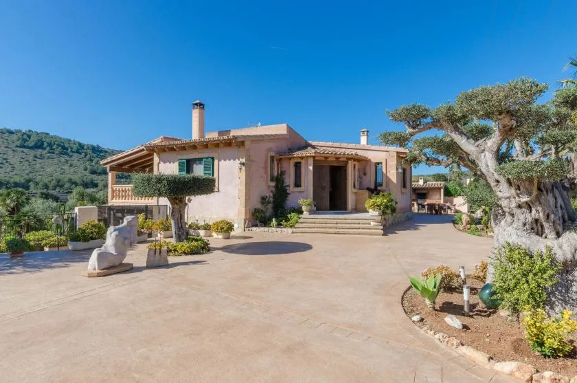 Una casa de estilo mediterráneo da a un amplio patio pavimentado con árboles y parterres.
