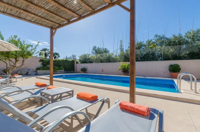 An outdoor swimming pool beside lounge seating under a shaded pergola.