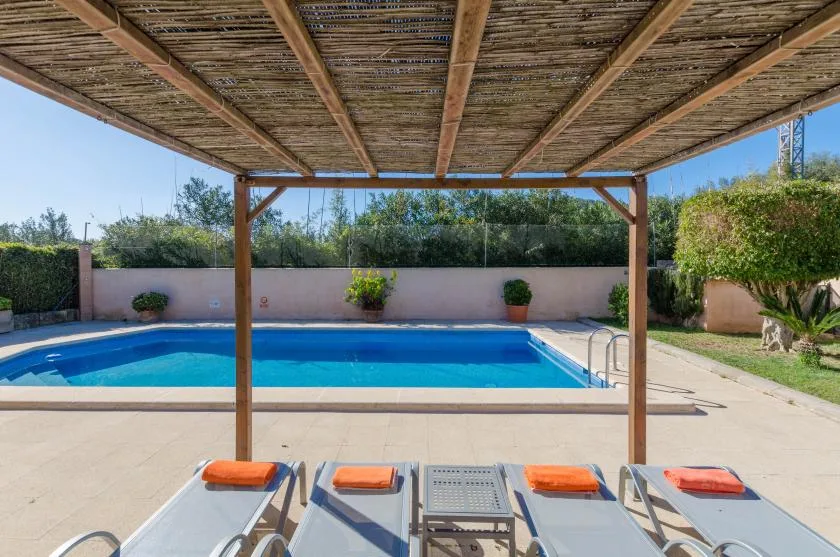 Una piscina está junto a tumbonas bajo una pérgola de madera con sombra.
