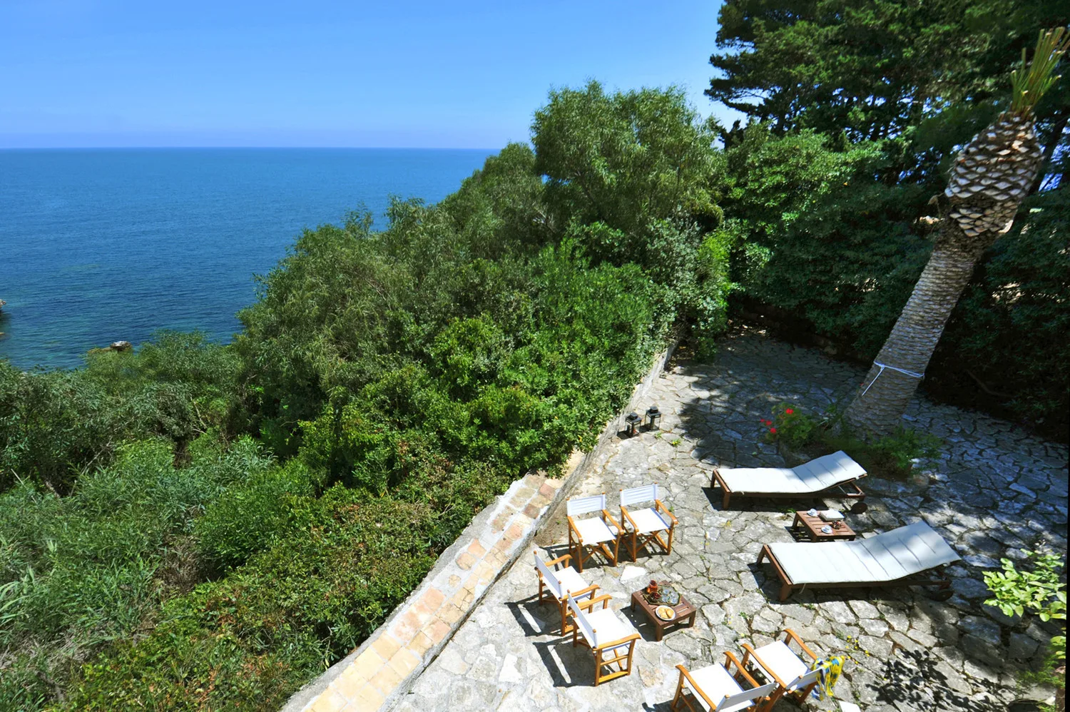 A stone patio with outdoor seating overlooks the sea and surrounding greenery.