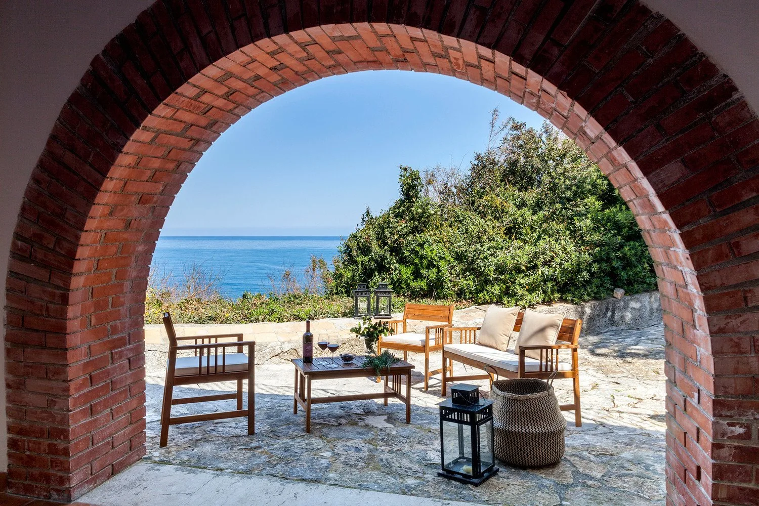 A stone patio with outdoor seating overlooks the sea through a brick arch.