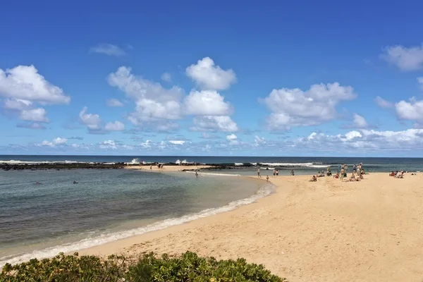 Una playa de arena se curva junto a aguas poco profundas bajo un cielo azul con nubes.