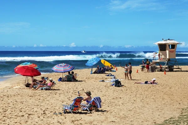 Una playa soleada con personas bajo sombrillas coloridas junto a las olas del océano.