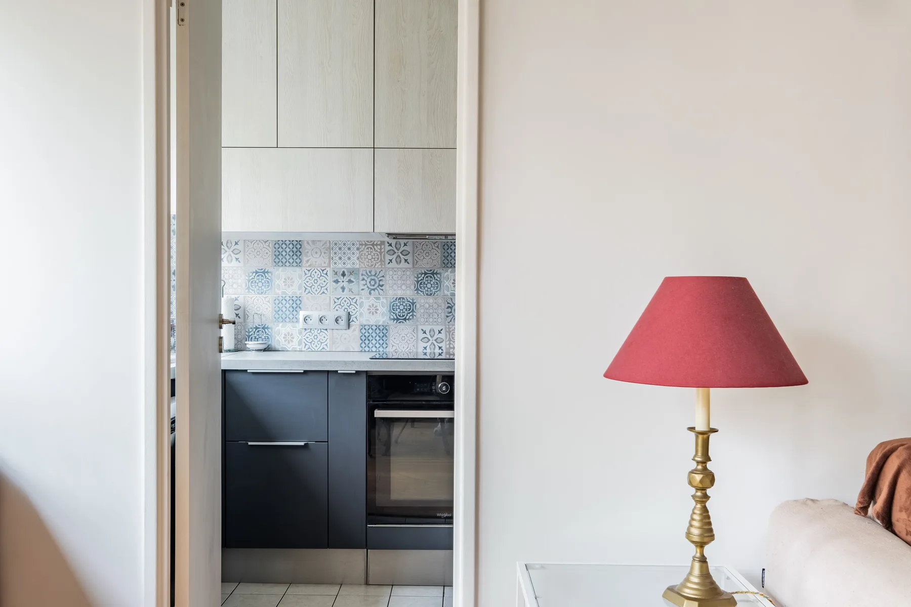 A compact kitchen is visible through a doorway beside a seating area with a red lamp.