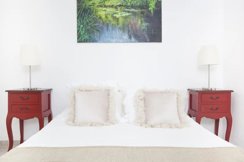 A bright bedroom with a white bed, red bedside tables, and a landscape painting.