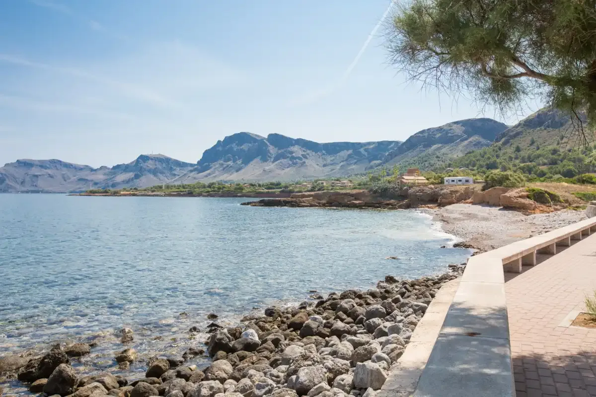 A rocky shoreline beside calm water, with a paved waterfront path and mountains beyond.