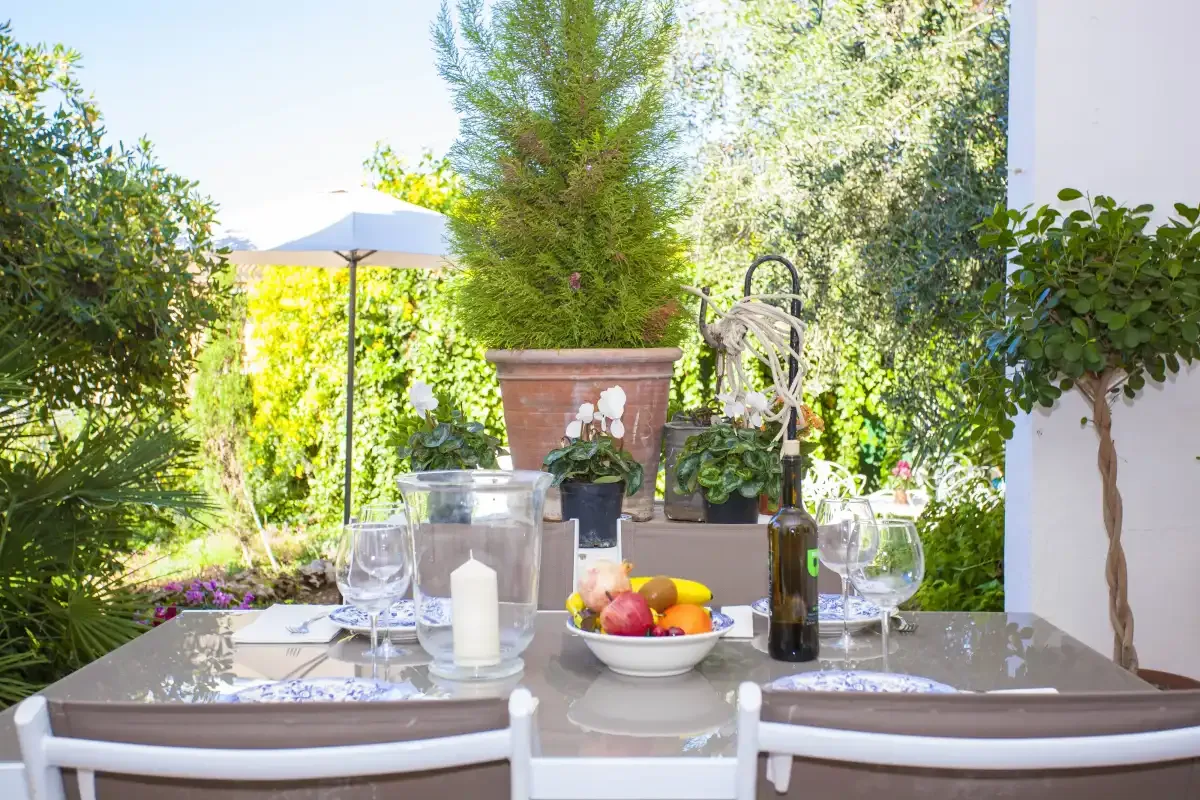 An outdoor dining table is set with glasses, fruit, and flowers in a leafy garden.