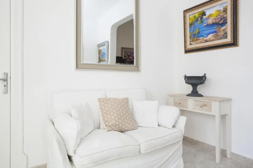 A bright living room with white seating beneath a wall mirror and framed painting.