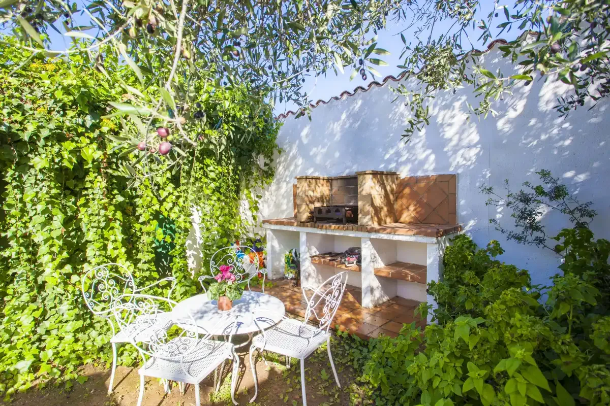 An outdoor patio with a built-in barbecue and metal seating beneath leafy branches.