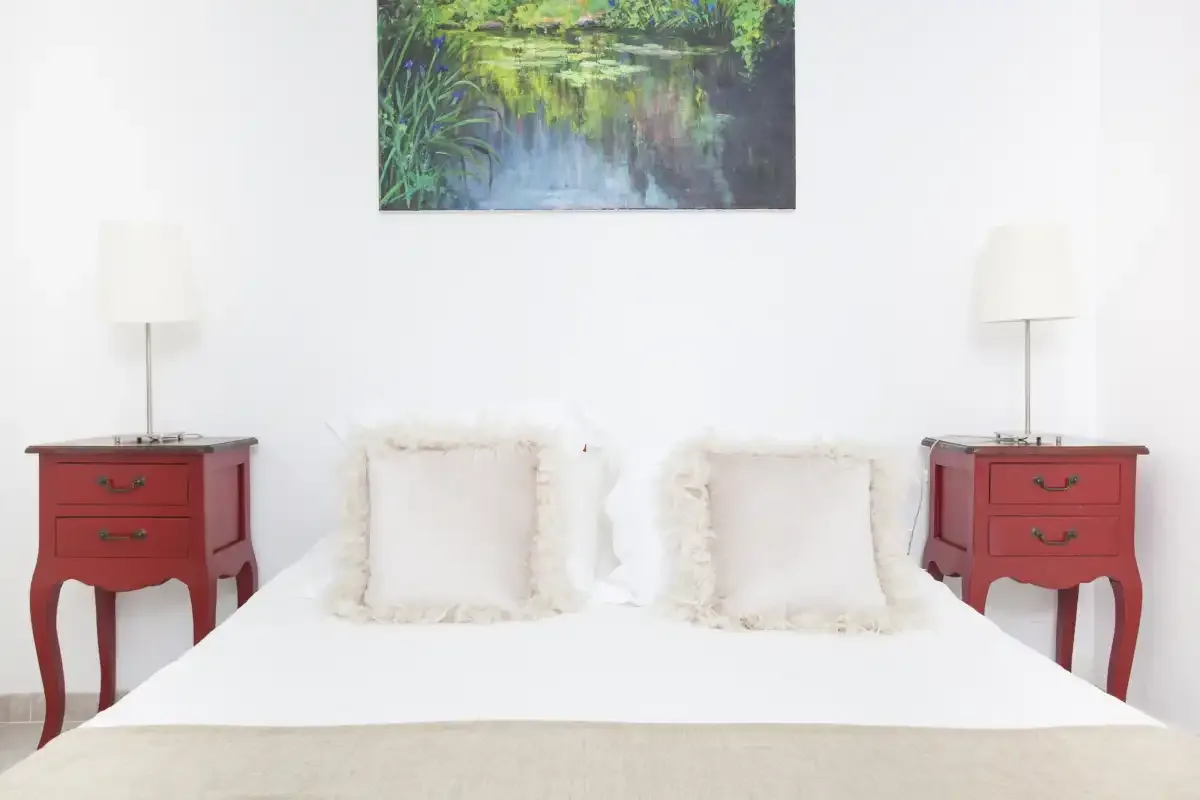 A bedroom with a white bed, red bedside tables, and a landscape painting.