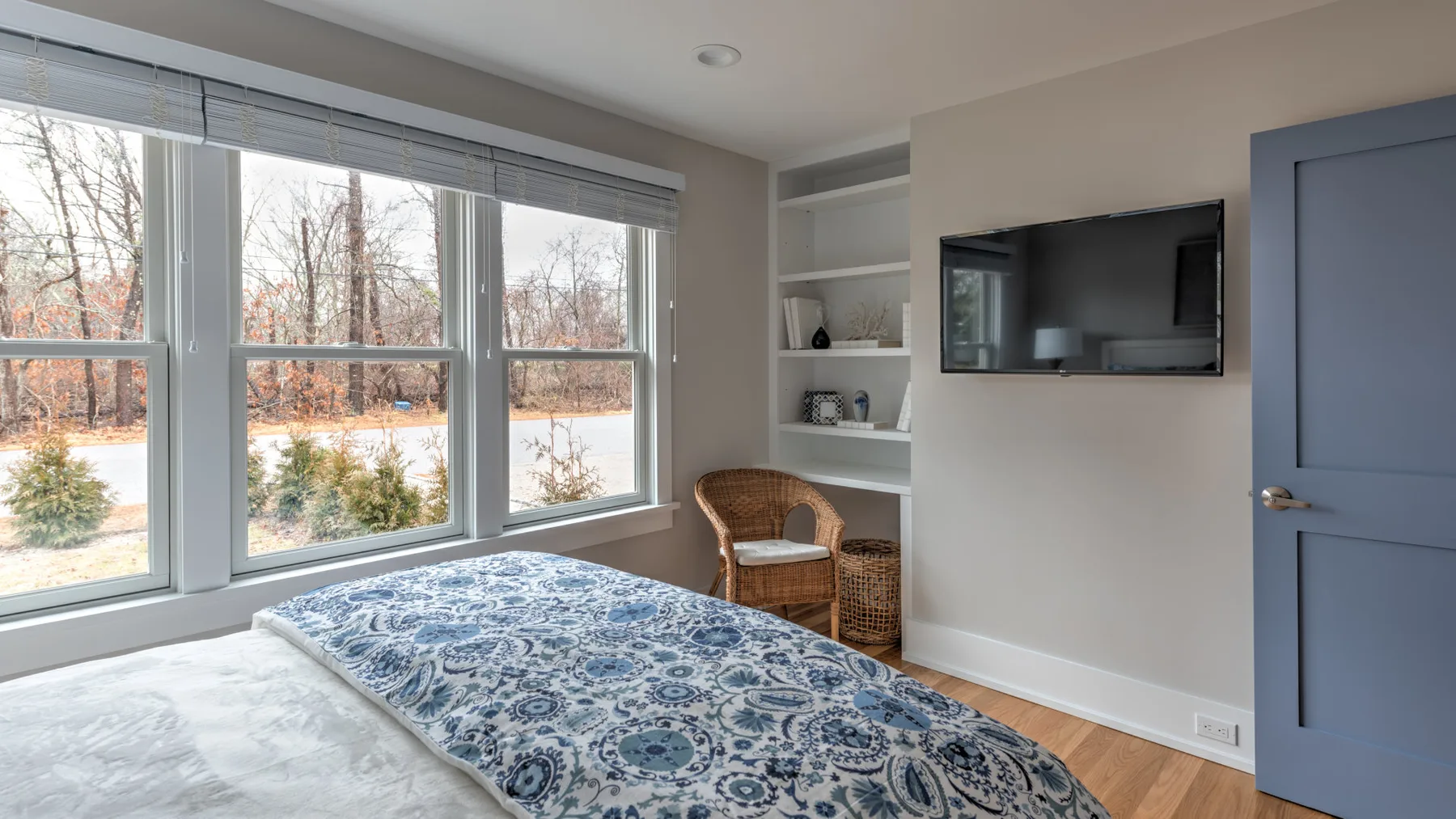 Un dormitorio con una colcha estampada junto a grandes ventanas y un televisor en la pared.