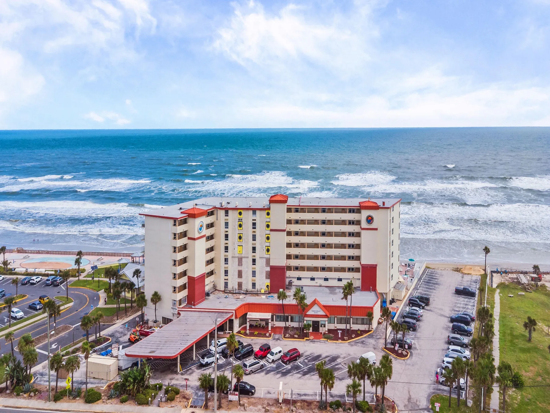 A beachfront hotel building stands beside the ocean, with a parking area in front.