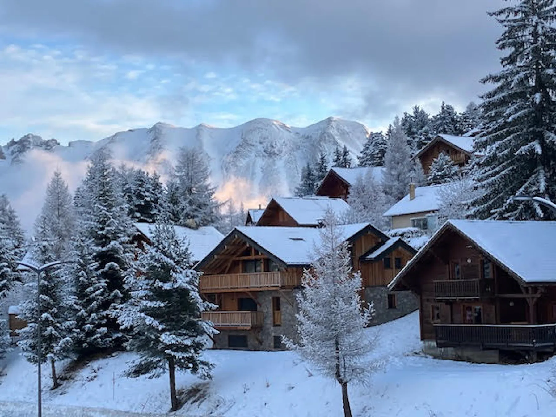 Snow-covered mountain chalets sit among evergreen trees beneath a cloudy sky.