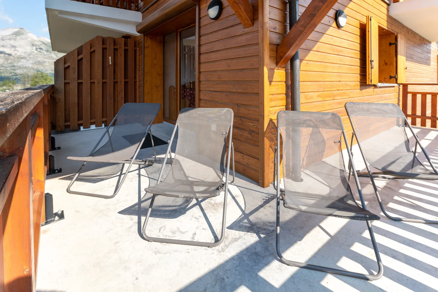 A sunny wooden balcony has several reclining seats and mountain views.