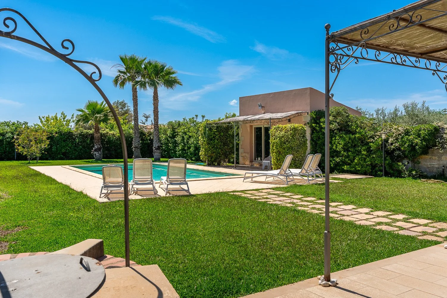 Zona de piscina al aire libre con asientos, palmeras y un patio con sombra.