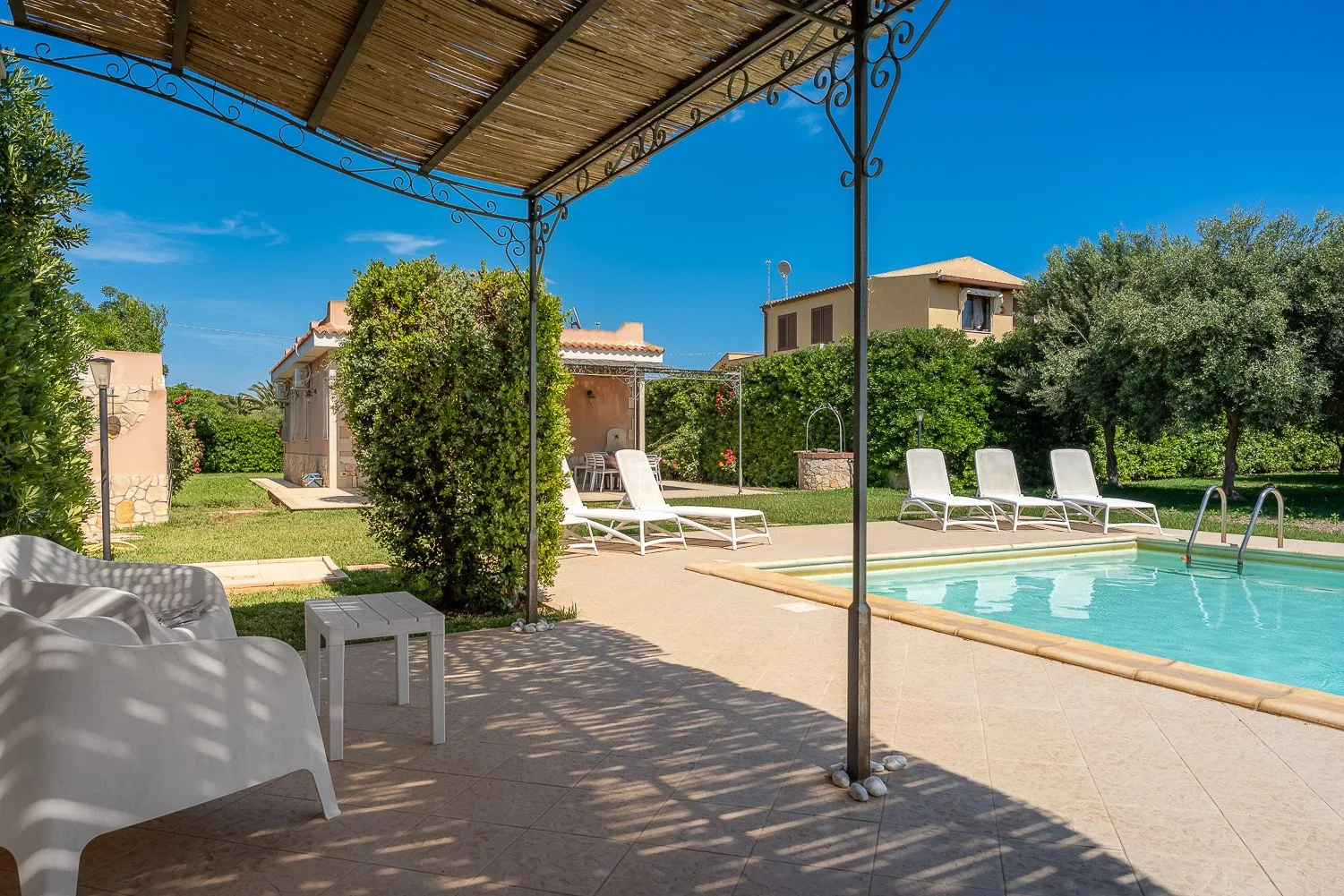 Una piscina junto a un patio ajardinado, con tumbonas bajo una pérgola sombreada.