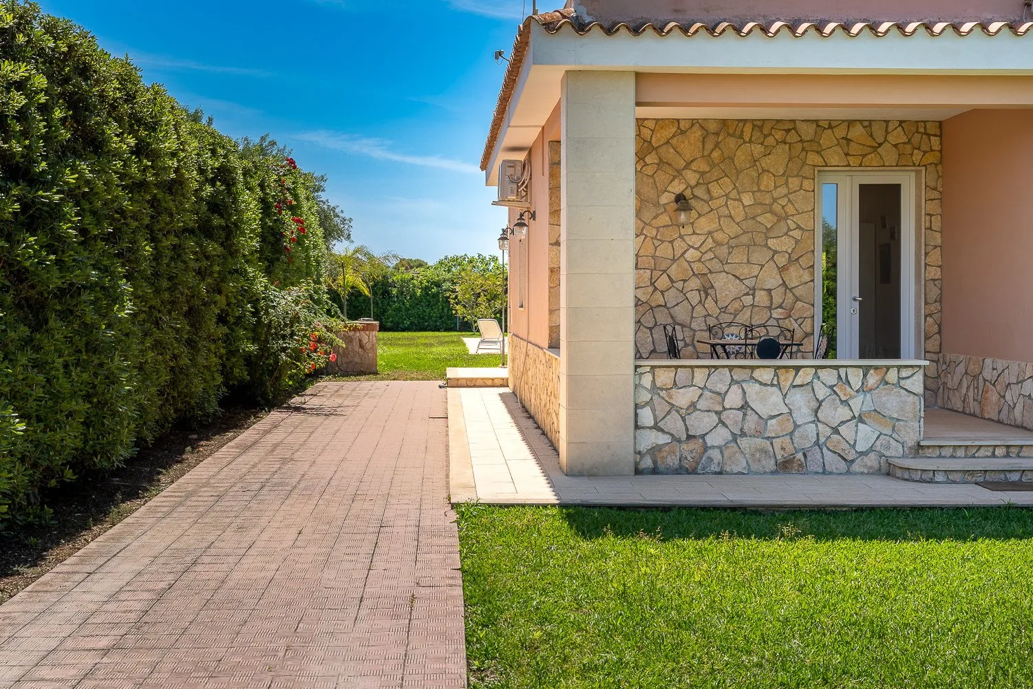 Un camino pavimentado pasa junto a una casa revestida de piedra y un césped verde.