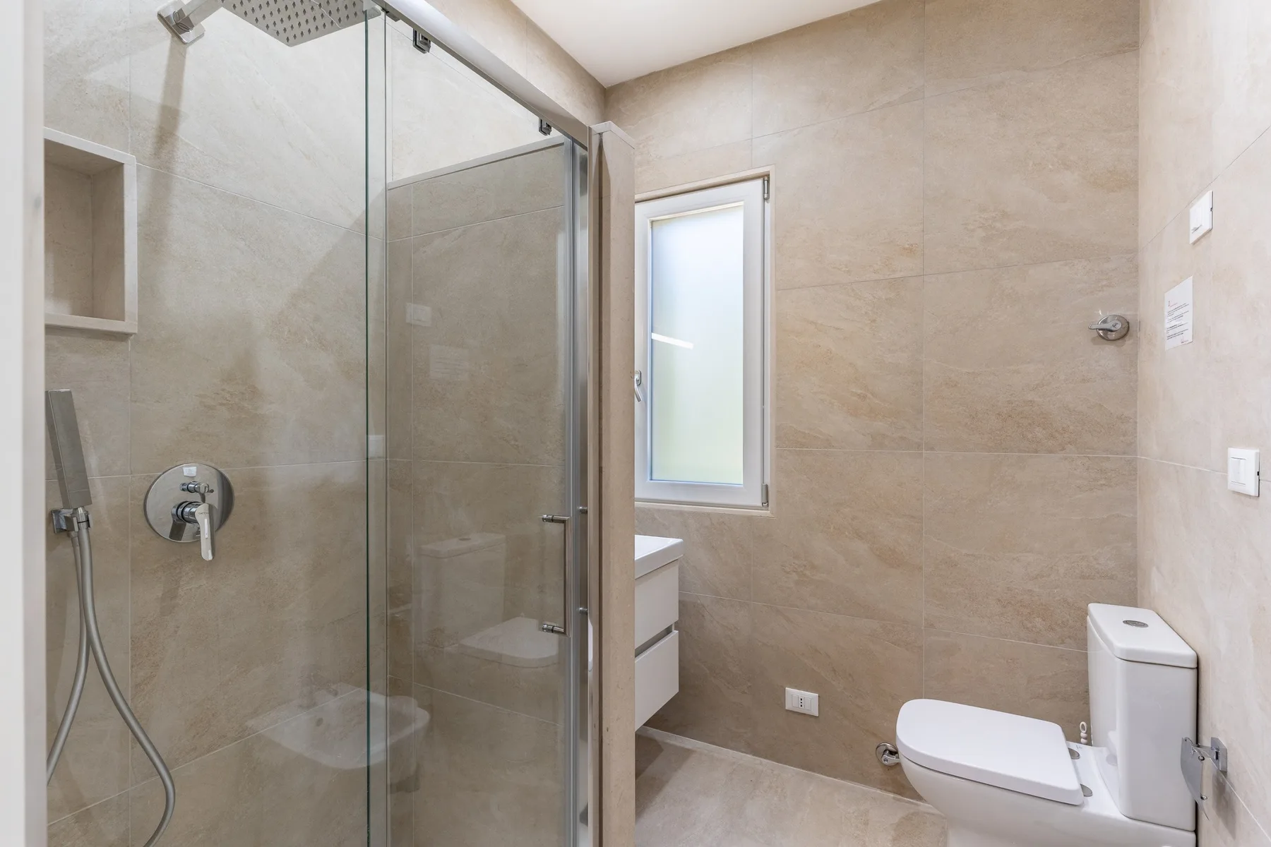 Baño alicatado con ducha de vidrio, ventana esmerilada e inodoro.