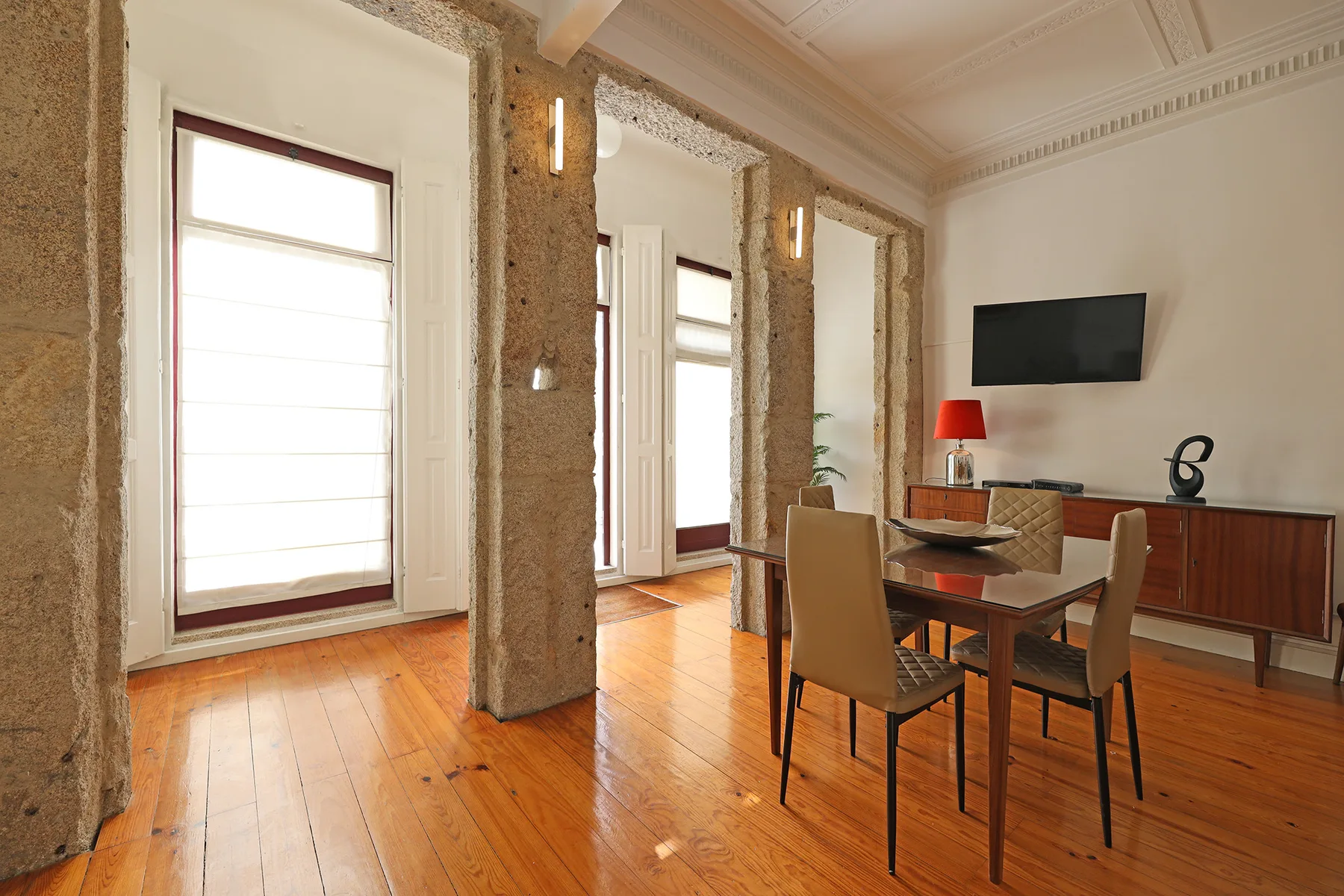 A dining area with a wooden table and chairs sits beside tall windows.