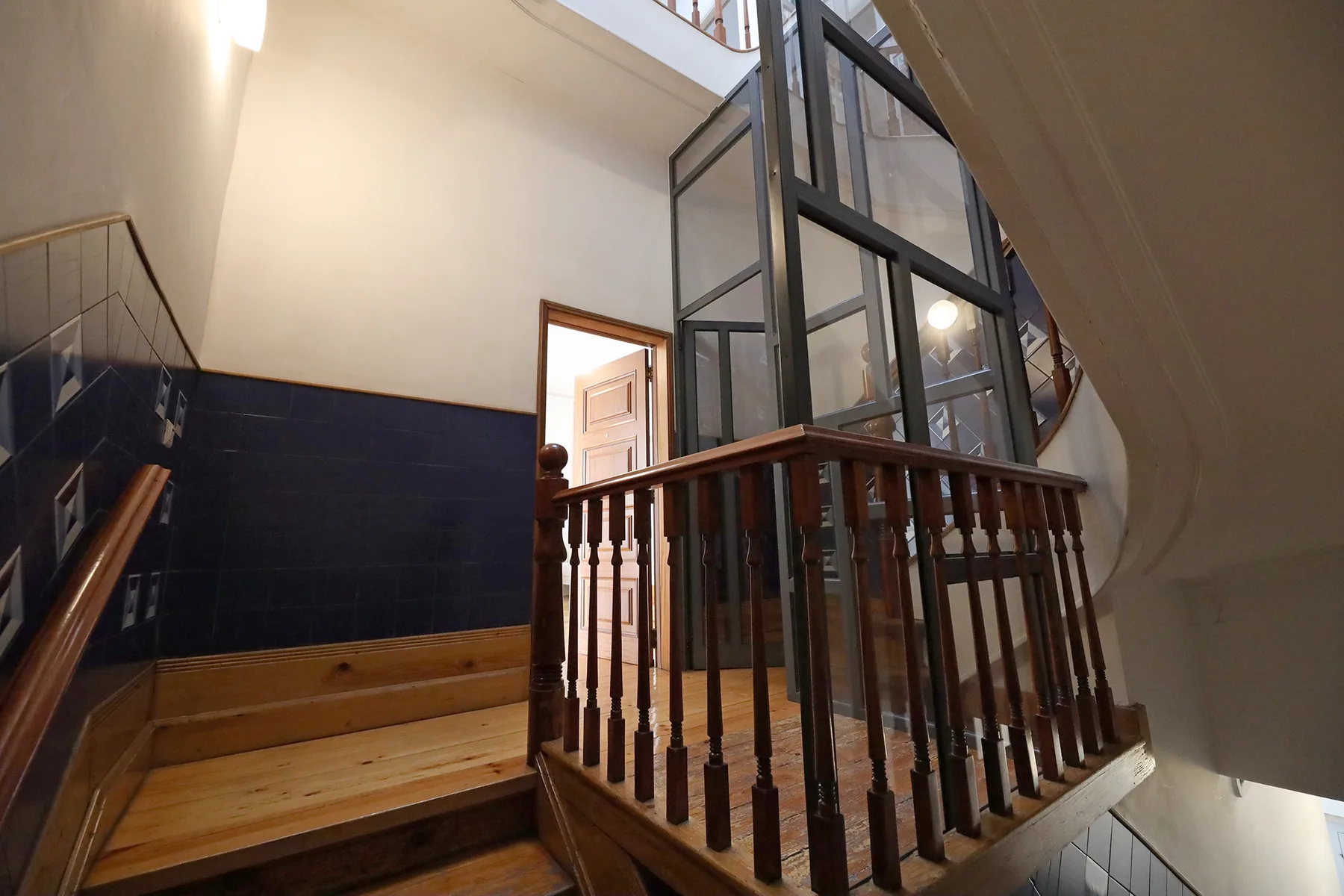 Una escalera interior con peldaños de madera y una cabina de ascensor acristalada.