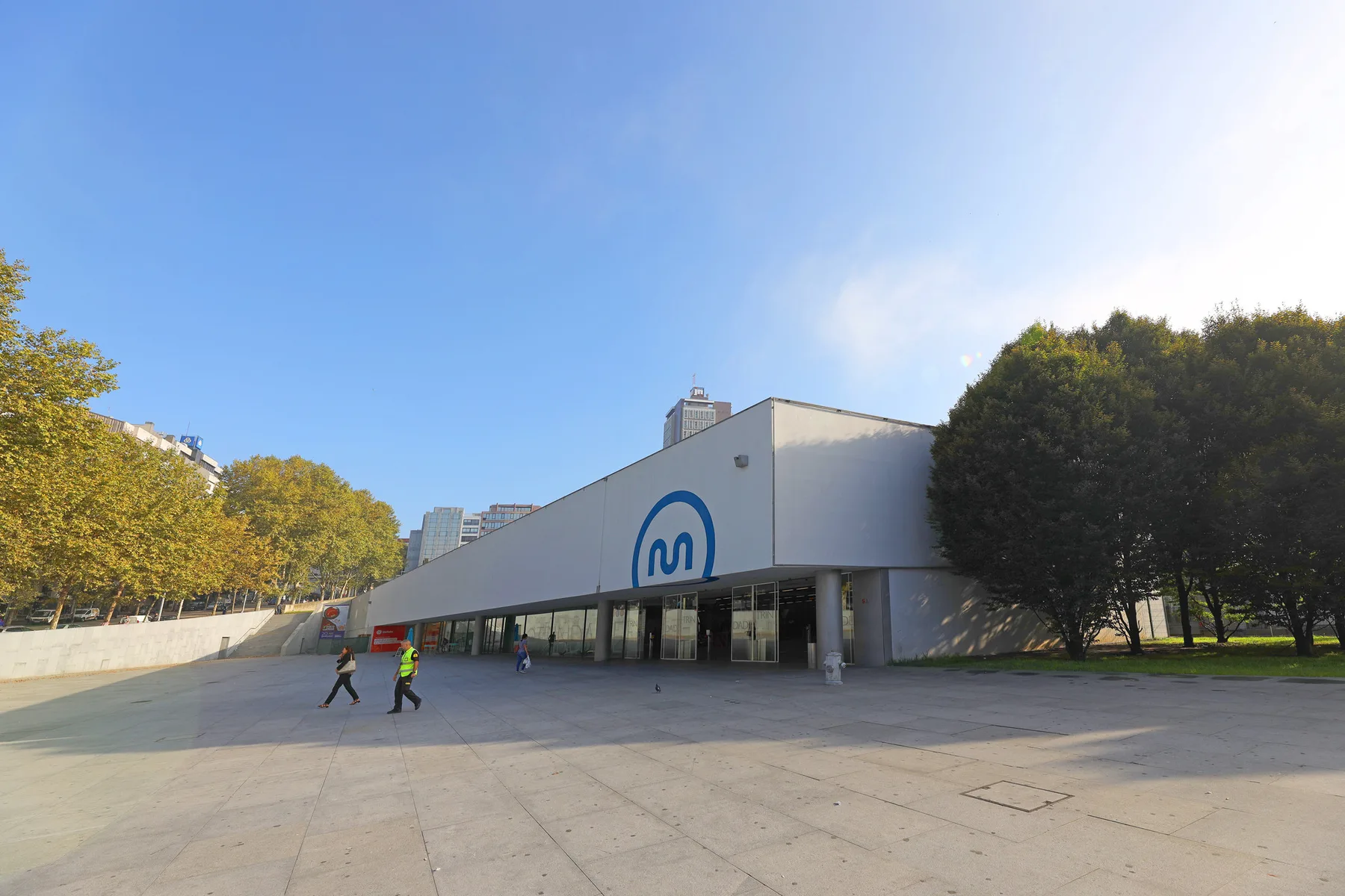 Un edificio público moderno junto a una amplia plaza pavimentada y árboles.