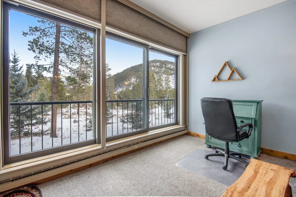 Un escritorio y una silla están junto a grandes ventanas con vistas a una ladera boscosa y nevada.